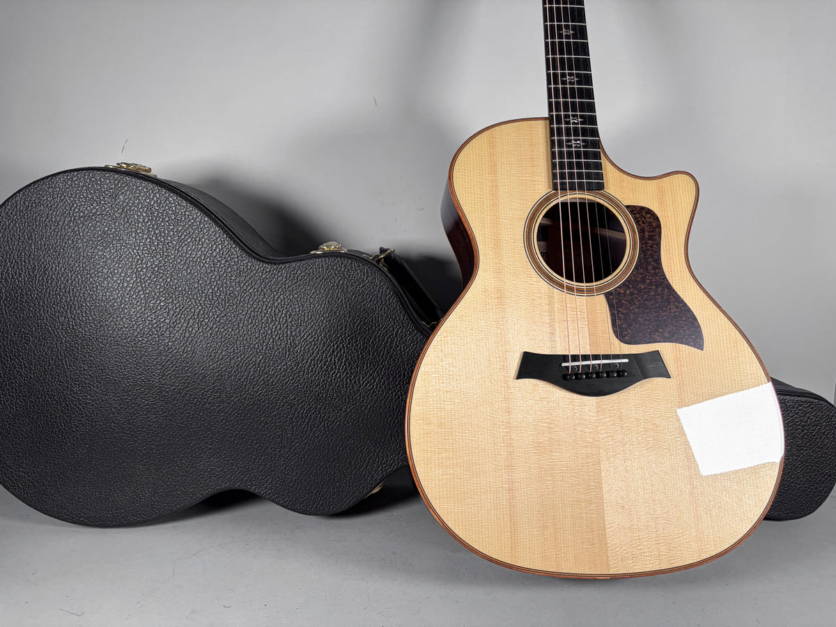 Light wood acoustic guitar with a cutaway body, dark pickguard, and black bridge, standing next to a black textured hard case.