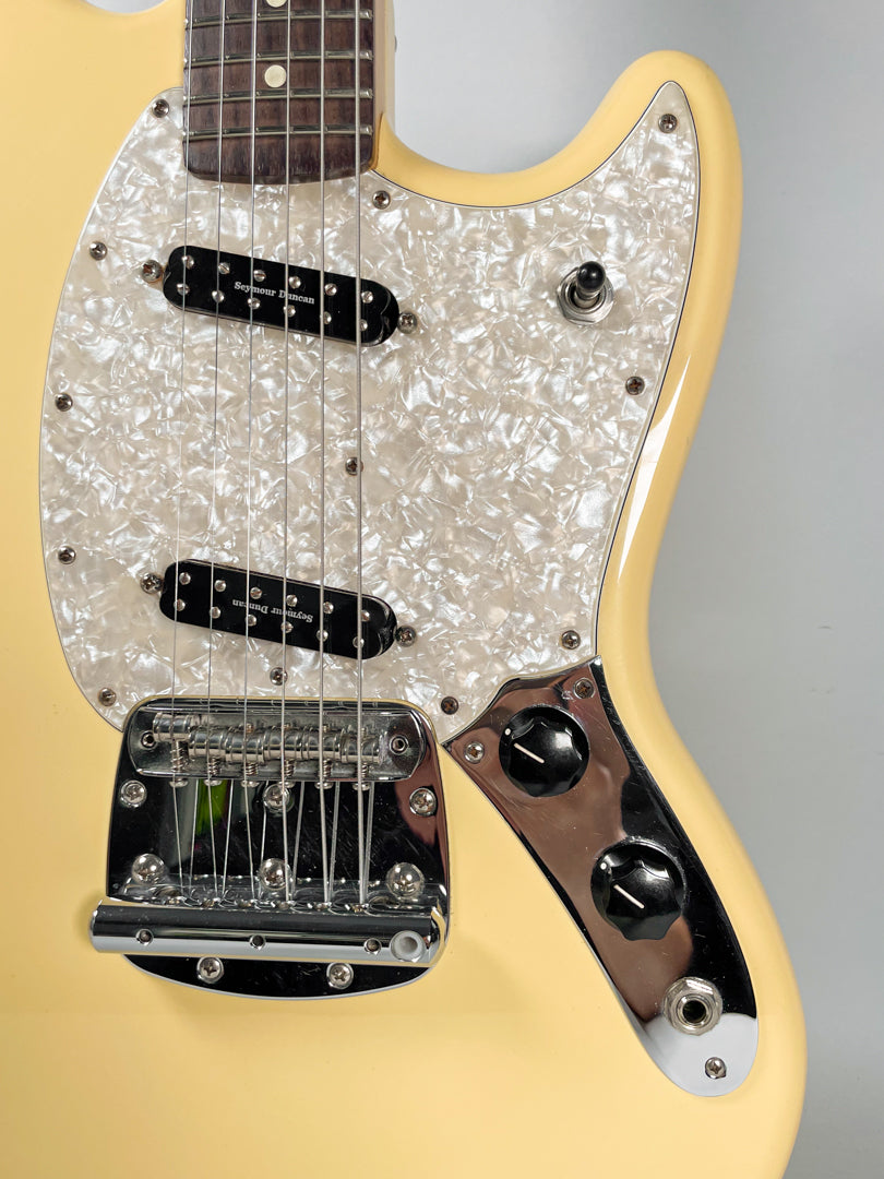 Close-up of a cream-colored electric guitar with a pearloid pickguard, two black pickups, a chrome bridge, and two black control knobs.