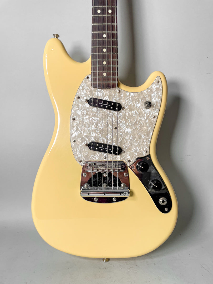 Cream-colored Mustang electric guitar, angled from above, showing its pearloid pickguard, two black pickups, and chrome bridge.