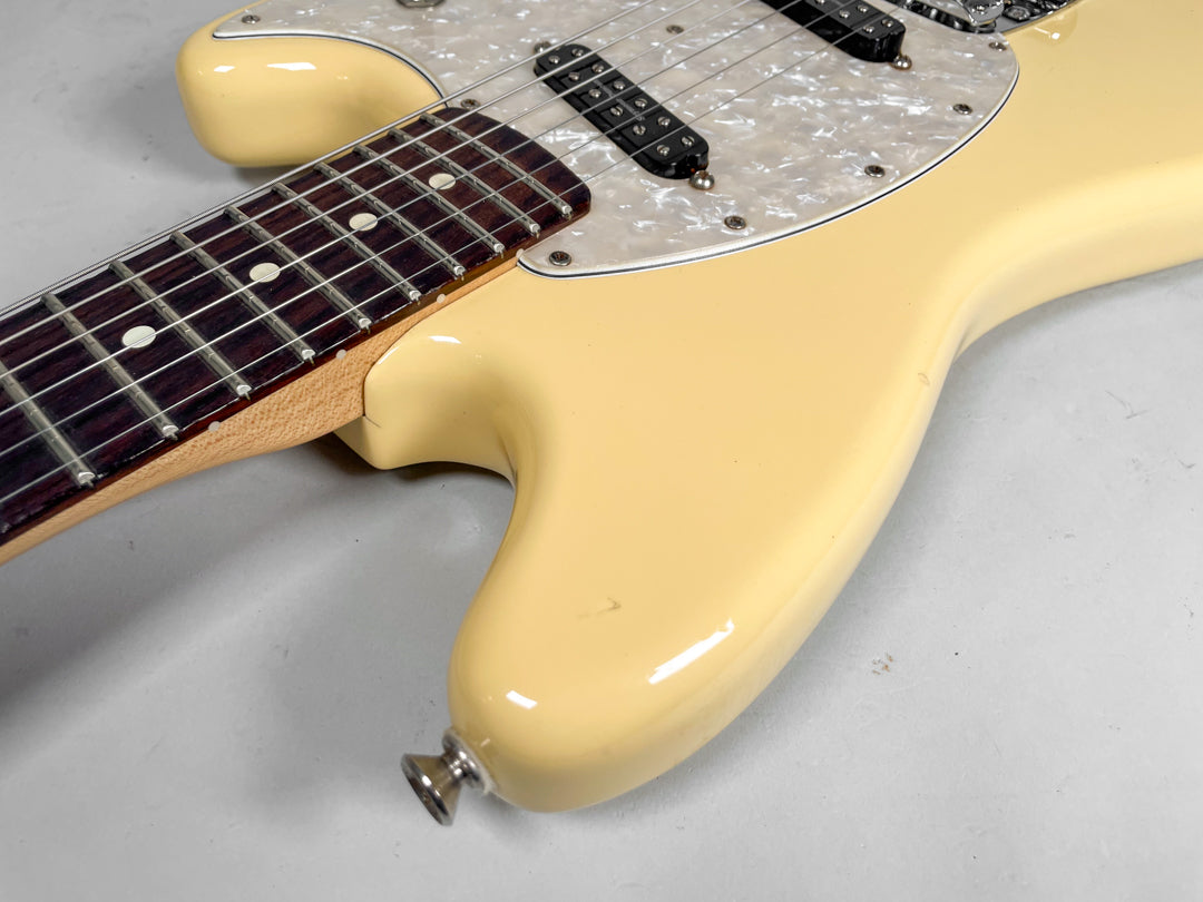 Close-up of a cream Mustang guitar body with a pearloid pickguard, dark fretboard, and two black pickups, showing a small ding.