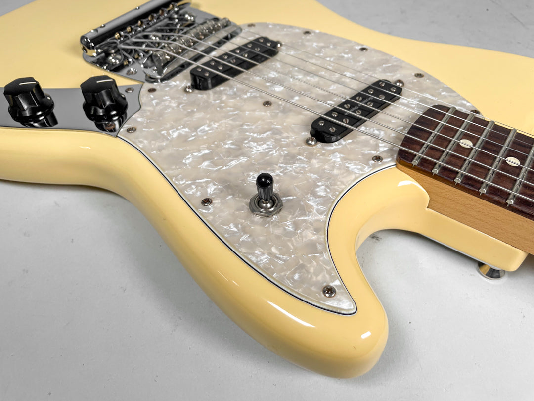 Close-up of a creamy yellow guitar body with a pearloid pickguard, two black knobs, a black toggle switch, and a chrome bridge.
