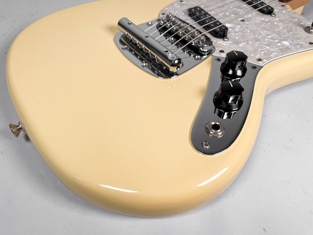 Close-up of the lower body of a cream Mustang guitar, showing the chrome bridge, pearloid pickguard, and two black control knobs.