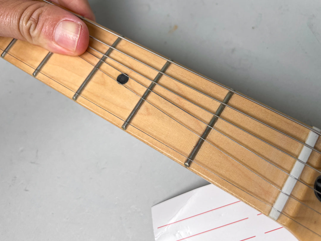 Close-up of a maple fretboard with black dot inlays, silver frets, and strings, held by a hand near the nut.