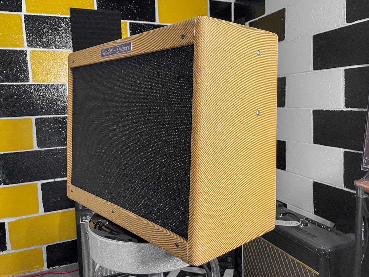 The tweed combo amp is angled, showing its textured tan finish, black grille cloth, and a blue "Fender - Deluxe" badge.