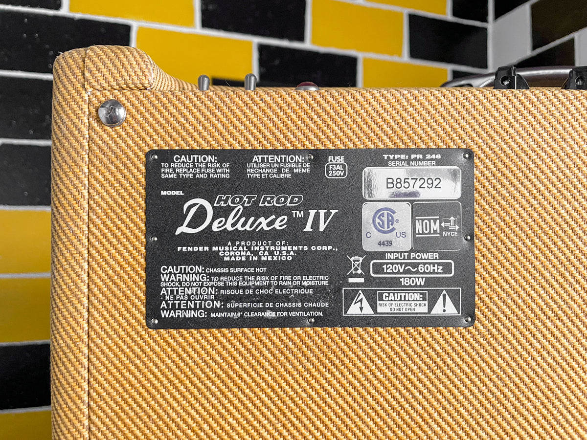 Close-up of the tweed-covered amp's rear panel, showing a black metal plate with white text and a visible serial number.