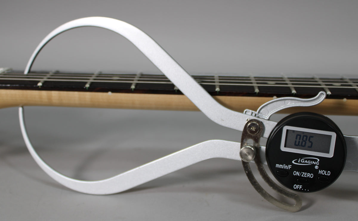 A digital caliper measures the neck thickness at the 1st fret, displaying 0.85 inches, with a maple neck and dark fretboard visible.