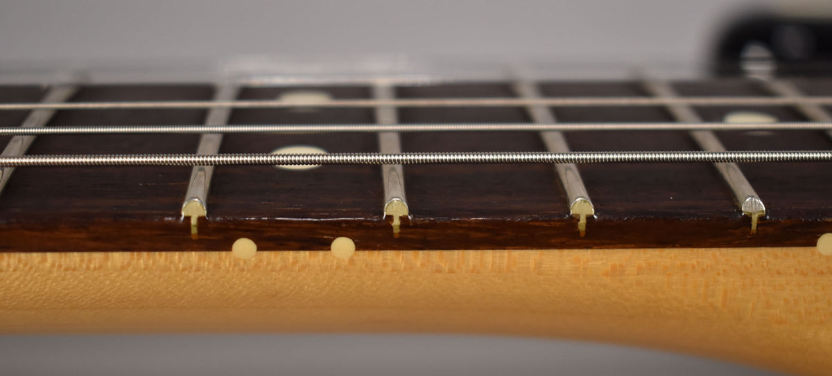 Close-up of the dark rosewood fretboard with white dot inlays, showing the frets and strings from a low angle, with a maple neck.