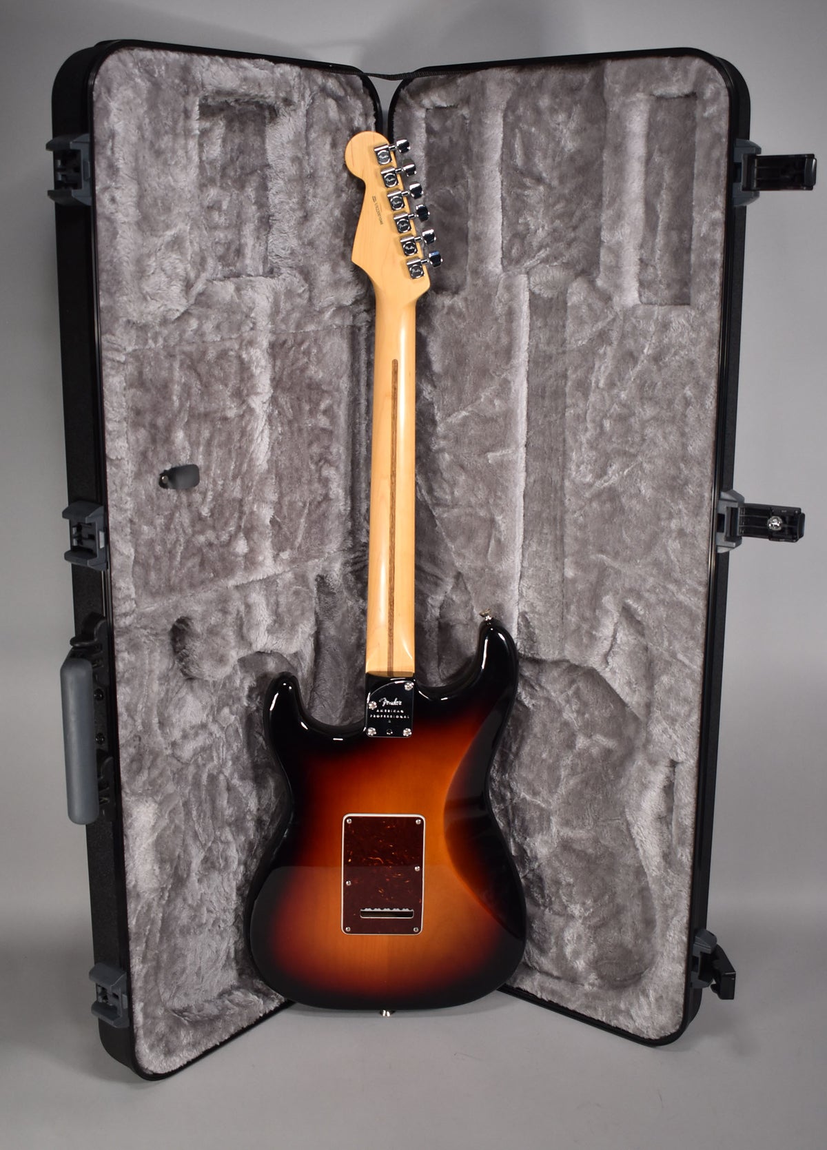 Rear view of a sunburst Stratocaster with a maple neck, chrome tuners, and a brown tremolo cavity cover, standing in its open case.