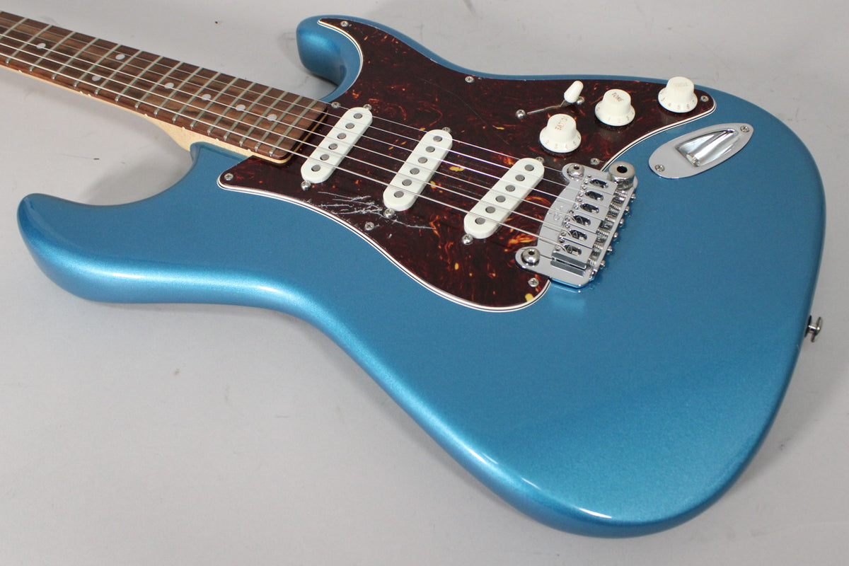 The metallic blue electric guitar is angled from the neck, showing its tortoiseshell pickguard, three white single-coil pickups, and chrome tremolo.