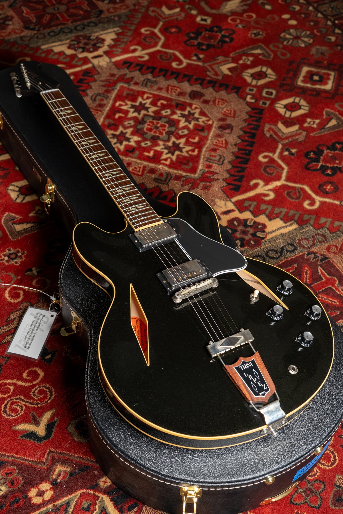 Black semi-hollowbody electric guitar with gold binding, f-holes, and a Trini Lopez tailpiece, resting in its open hard case on a red rug.