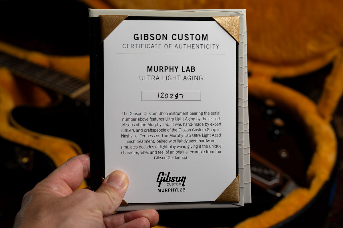 A hand holds the guitar's white Certificate of Authenticity with gold corners, partially obscuring the guitar in its brown case.