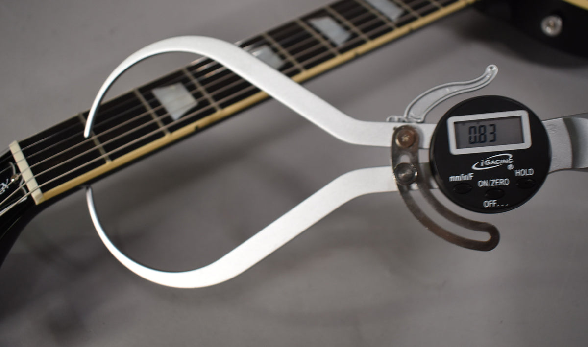 A digital caliper measures the string height at the nut of the guitar, showing 0.83mm.