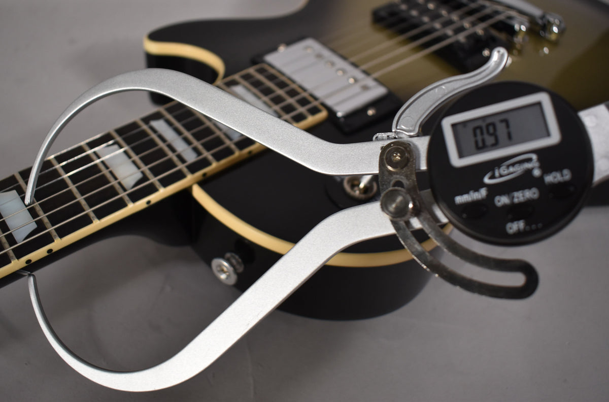 A digital caliper measures the guitar's nut width at 0.97 inches, with the black body and cream binding visible behind it.