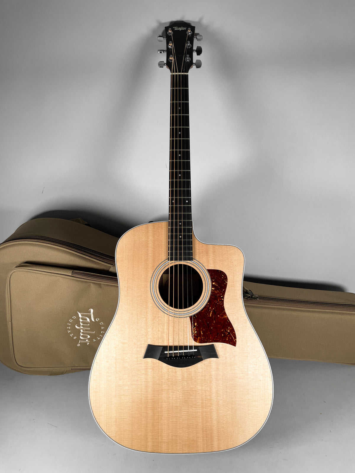 Full-front view of a natural finish acoustic guitar with a cutaway body, tortoise pickguard, and a tan soft case behind it.