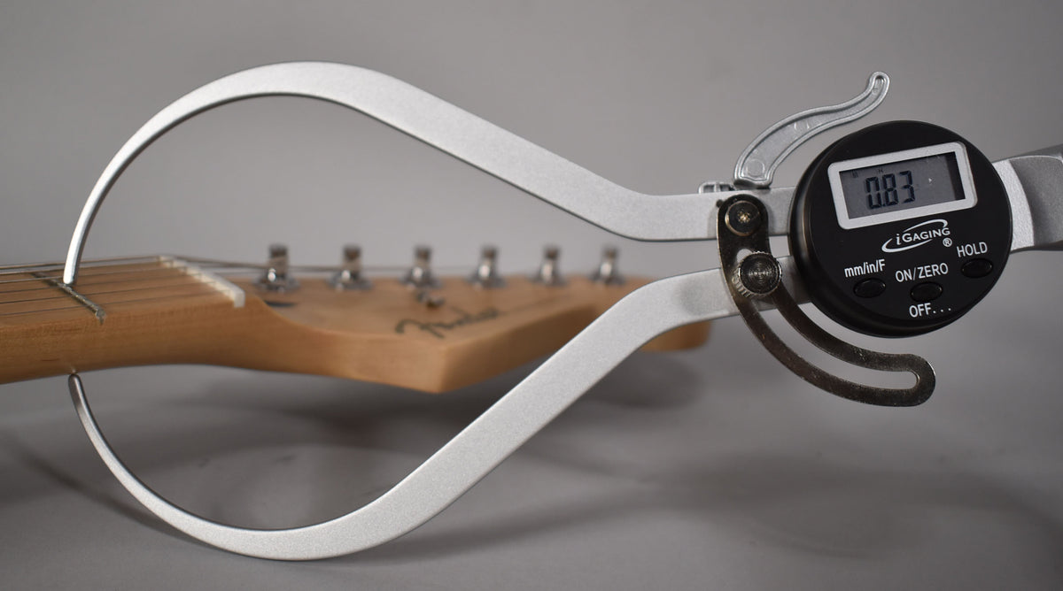 A digital caliper measures the string height at the first fret of the Stratocaster's light wood neck, displaying "0.83".