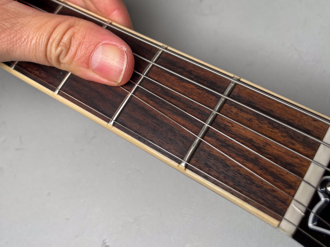 Close-up of a hand fretting the dark rosewood fingerboard with cream binding, showing the nut and first few frets.