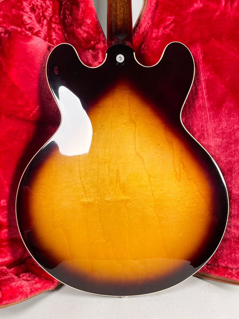 The guitar's sunburst back, with a glossy finish and visible wood grain, rests in a red velvet case, showing no wear.