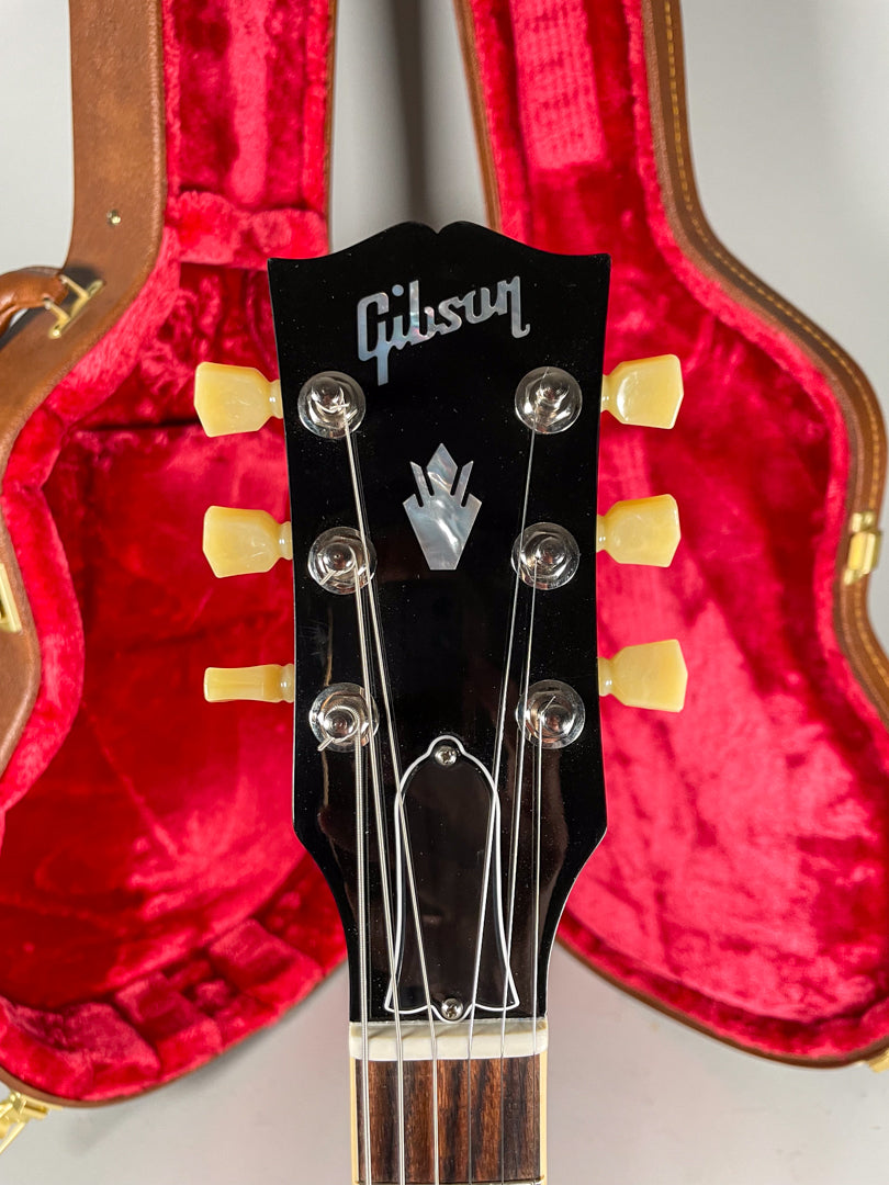 The guitar's glossy black headstock with mother-of-pearl inlay and cream tuning pegs is framed against a red velvet-lined case.