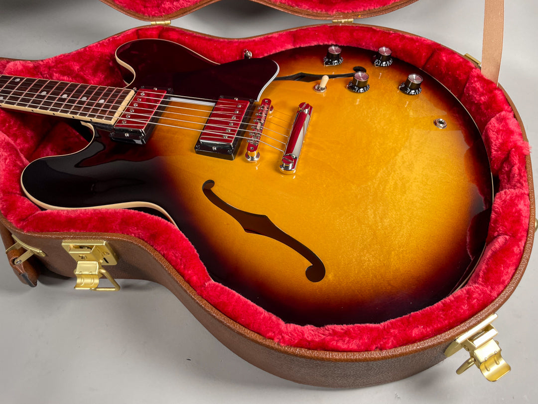 The sunburst electric guitar rests in its red-lined brown case, showing its f-hole, chrome hardware, and four black control knobs.