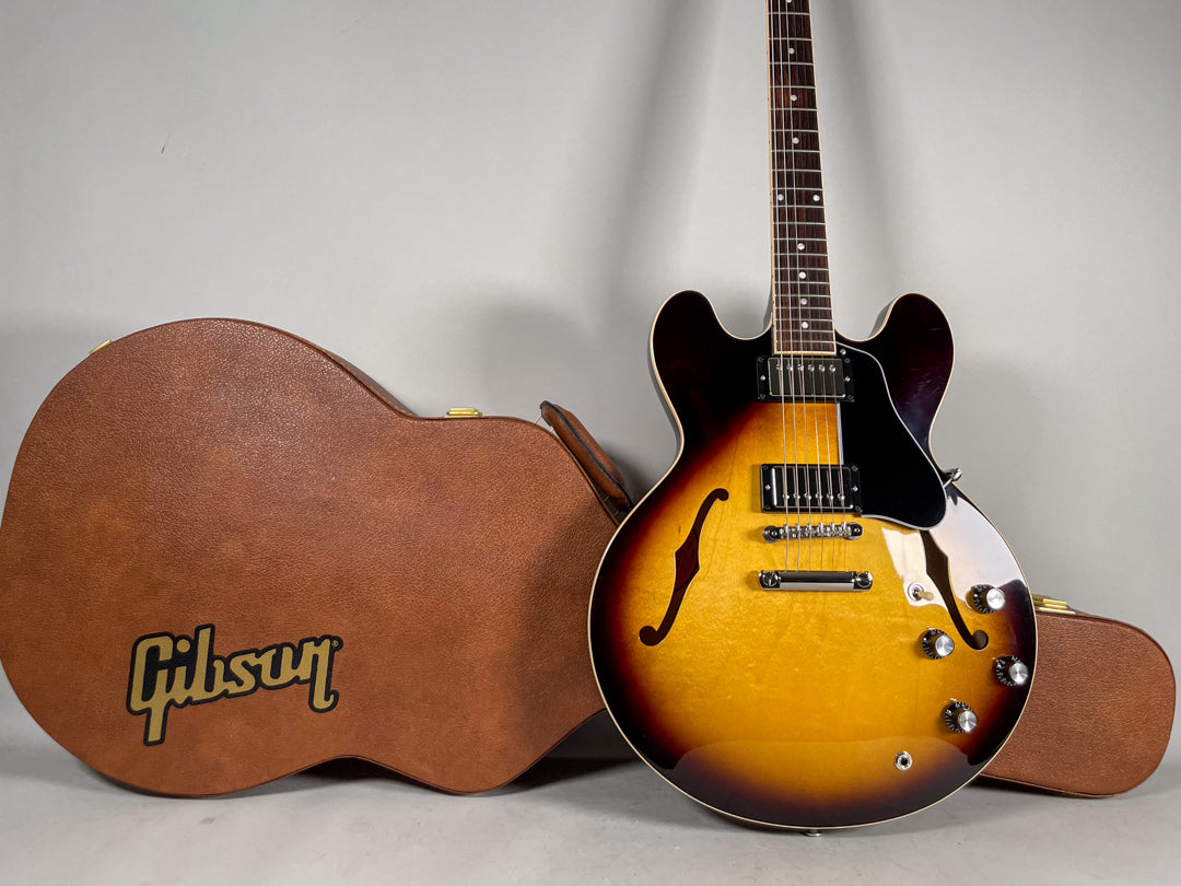 Sunburst semi-hollow electric guitar with black humbuckers, chrome hardware, and four black control knobs, next to a brown hard case.
