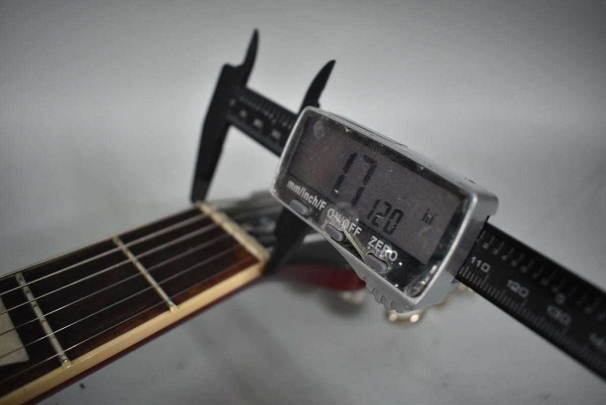 Close-up of the guitar's dark fretboard and strings, with a digital caliper measuring the nut width at 1.712 inches.
