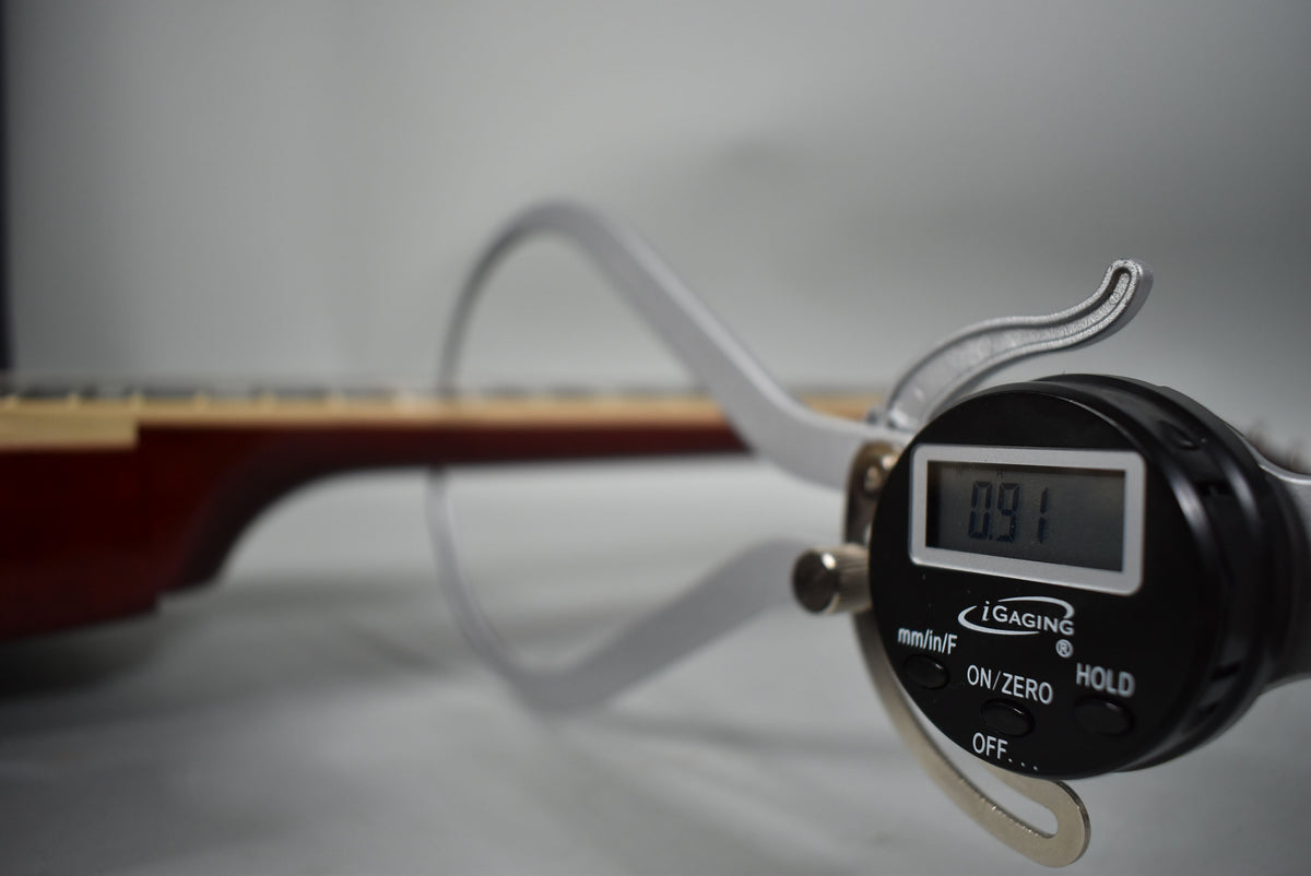 A digital caliper measures the guitar's neck thickness at 0.91 inches, with the sunburst body blurred in the background.