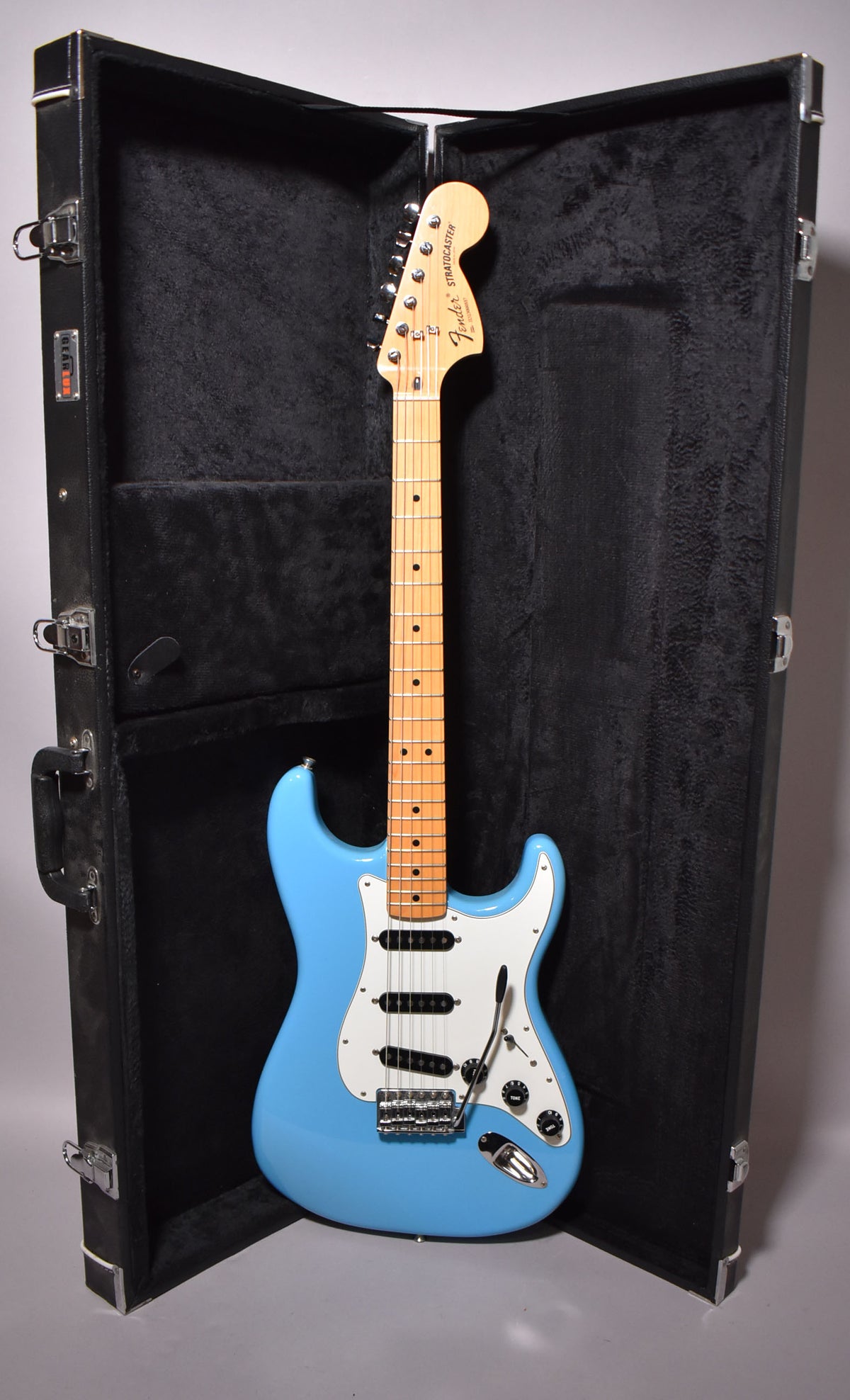 Full-length view of a light blue Stratocaster with a maple neck and white pickguard, standing upright in its open black hard case.