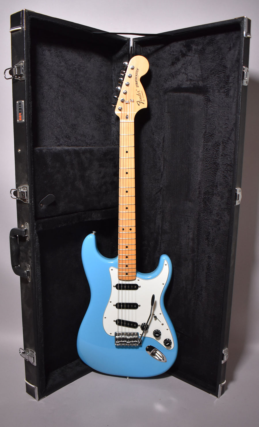 Full-length view of a light blue Stratocaster with a maple neck and white pickguard, standing upright in its open black hard case.