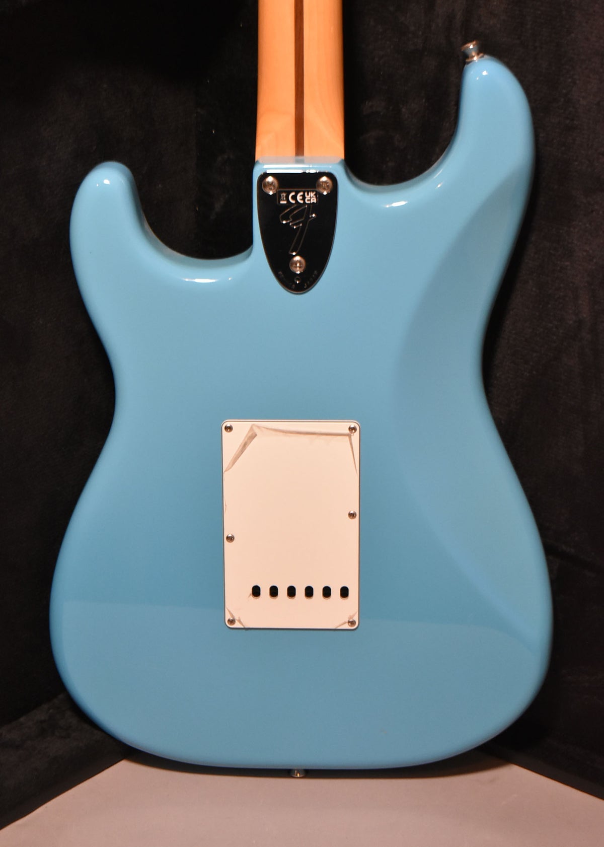 The back of a glossy Maui Blue Stratocaster, showing the neck plate, a light maple neck, and a white tremolo cover with peeling plastic film.