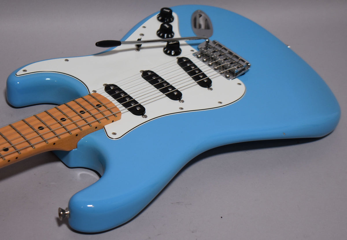 Light blue electric guitar, angled from above, showing the body, white pickguard, three black single-coil pickups, and maple fretboard.