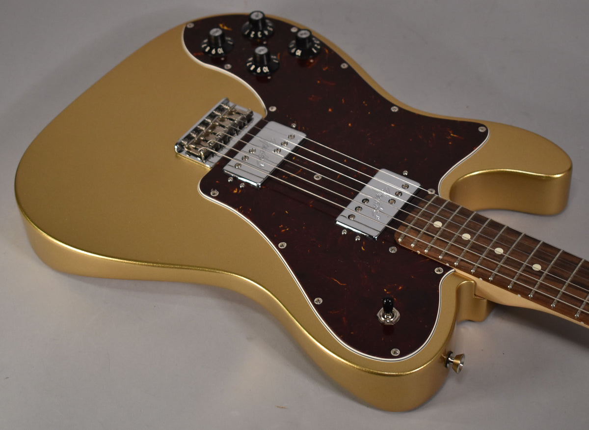 The gold left-handed Telecaster is angled from the upper left, revealing its tortoiseshell pickguard, two chrome humbuckers, and four black control knobs.
