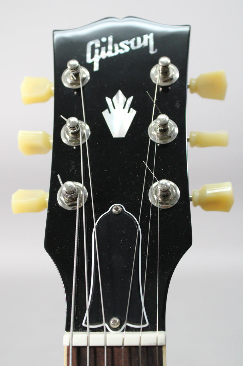 Close-up of the guitar's glossy black headstock with cream tuning pegs, a mother-of-pearl crown inlay, and a bell-shaped truss rod cover.