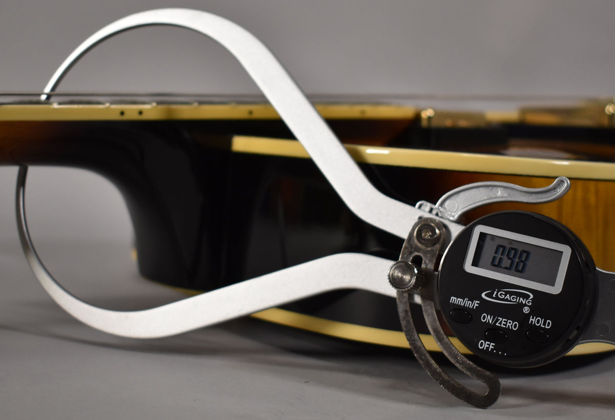 A digital caliper measures the guitar's neck thickness at 0.98, showing the dark sunburst finish and cream binding.