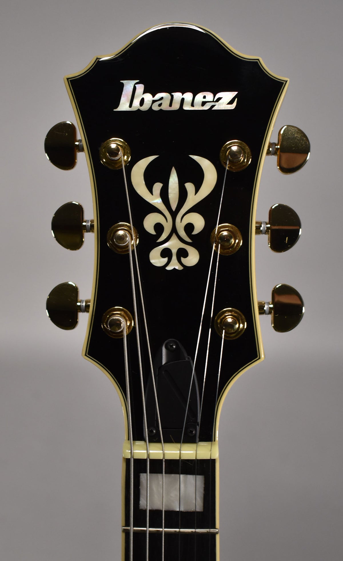 The guitar's black headstock with cream binding and gold tuners is shown, featuring a mother-of-pearl Ibanez logo and ornate inlay.