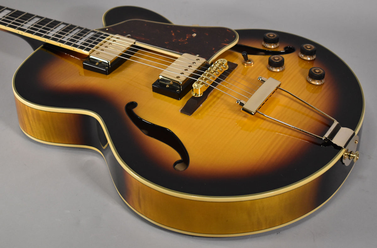 Close-up of a sunburst archtop guitar, angled from the upper left, showing the body, f-holes, gold hardware, and four control knobs.