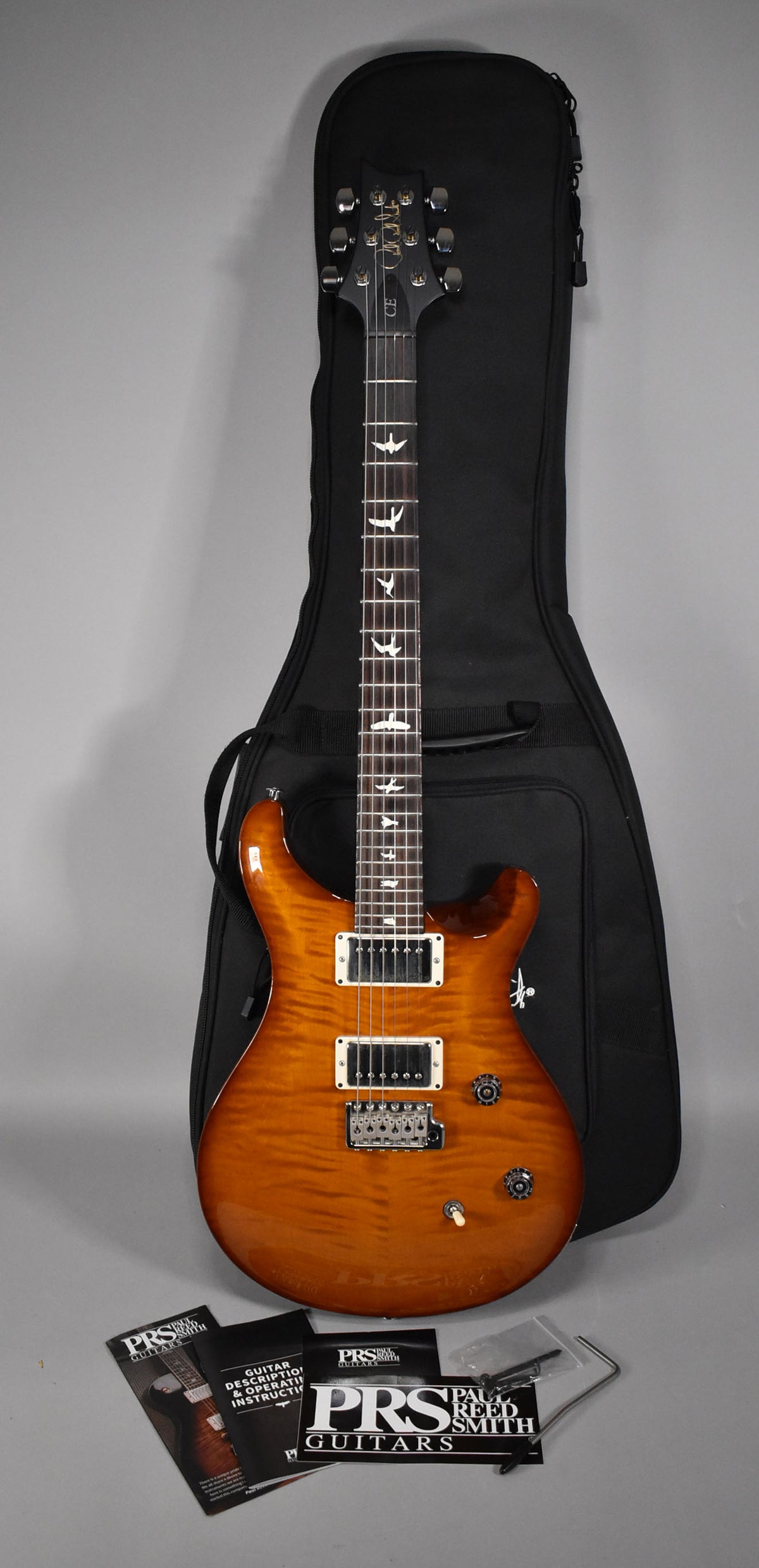 Full-length view of a flamed maple top electric guitar in a tobacco burst finish, with a black gig bag behind it.