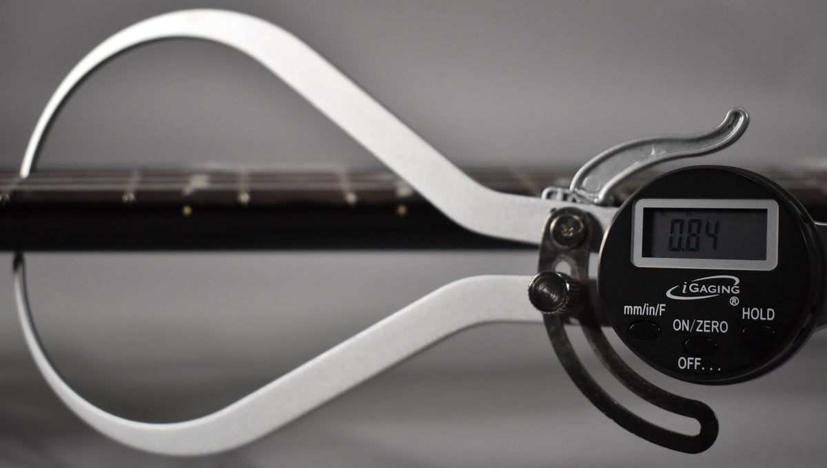 A digital caliper measures the guitar's fretboard, displaying "0.84" on its screen, with the dark fretboard and strings visible.