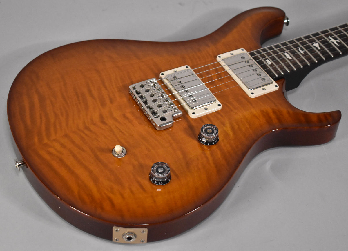 The electric guitar's flame maple top in a warm amber burst finish, with chrome hardware and cream pickup rings, is angled from the lower bout.