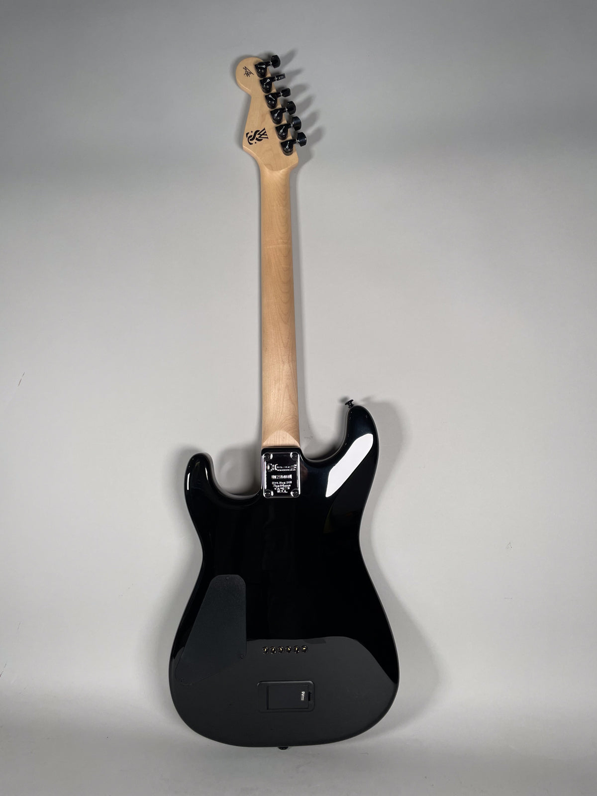 The back of a black electric guitar with a glossy finish, a light wood neck, and black hardware, including a battery compartment.