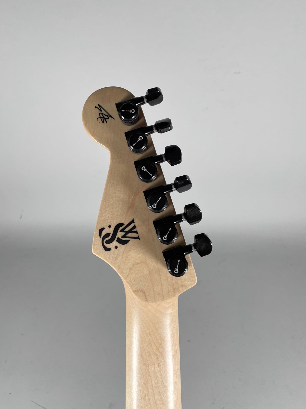 Rear view of the guitar's natural wood headstock and neck, showing six black tuning pegs and two black graphic logos.