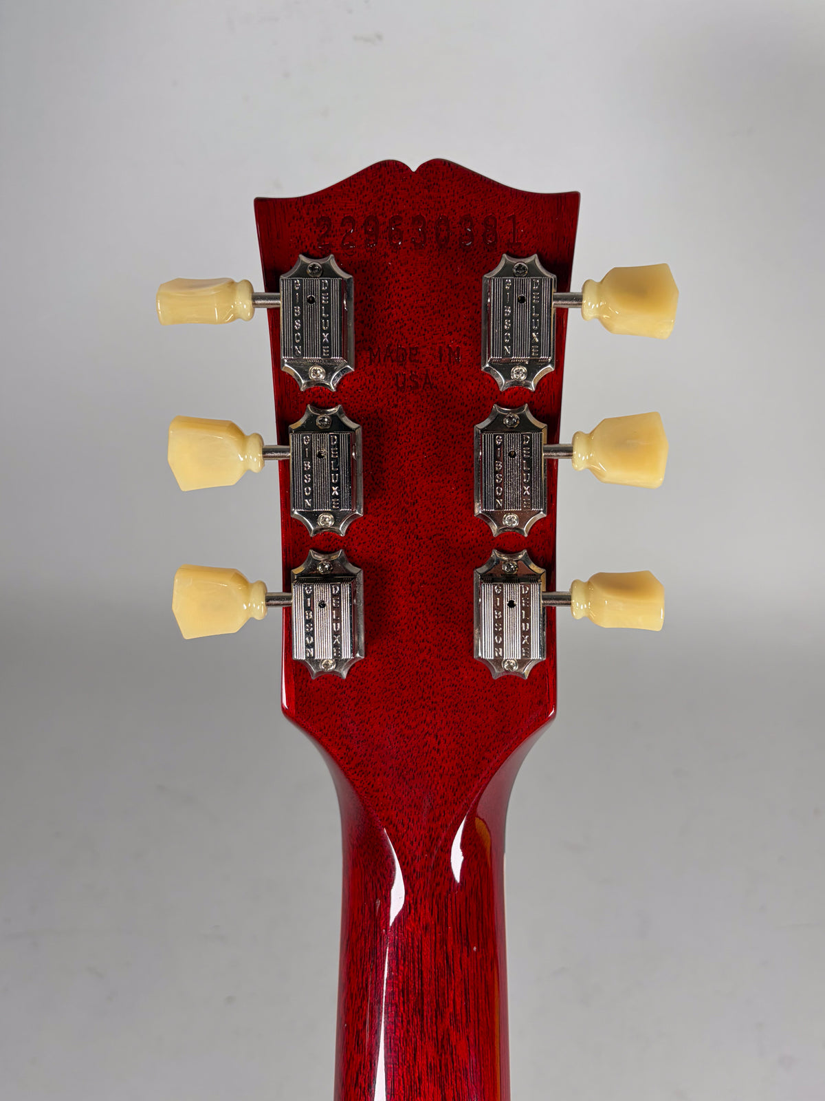 The back of a cherry red headstock with six cream tuning pegs and chrome tuners, showing a serial number.