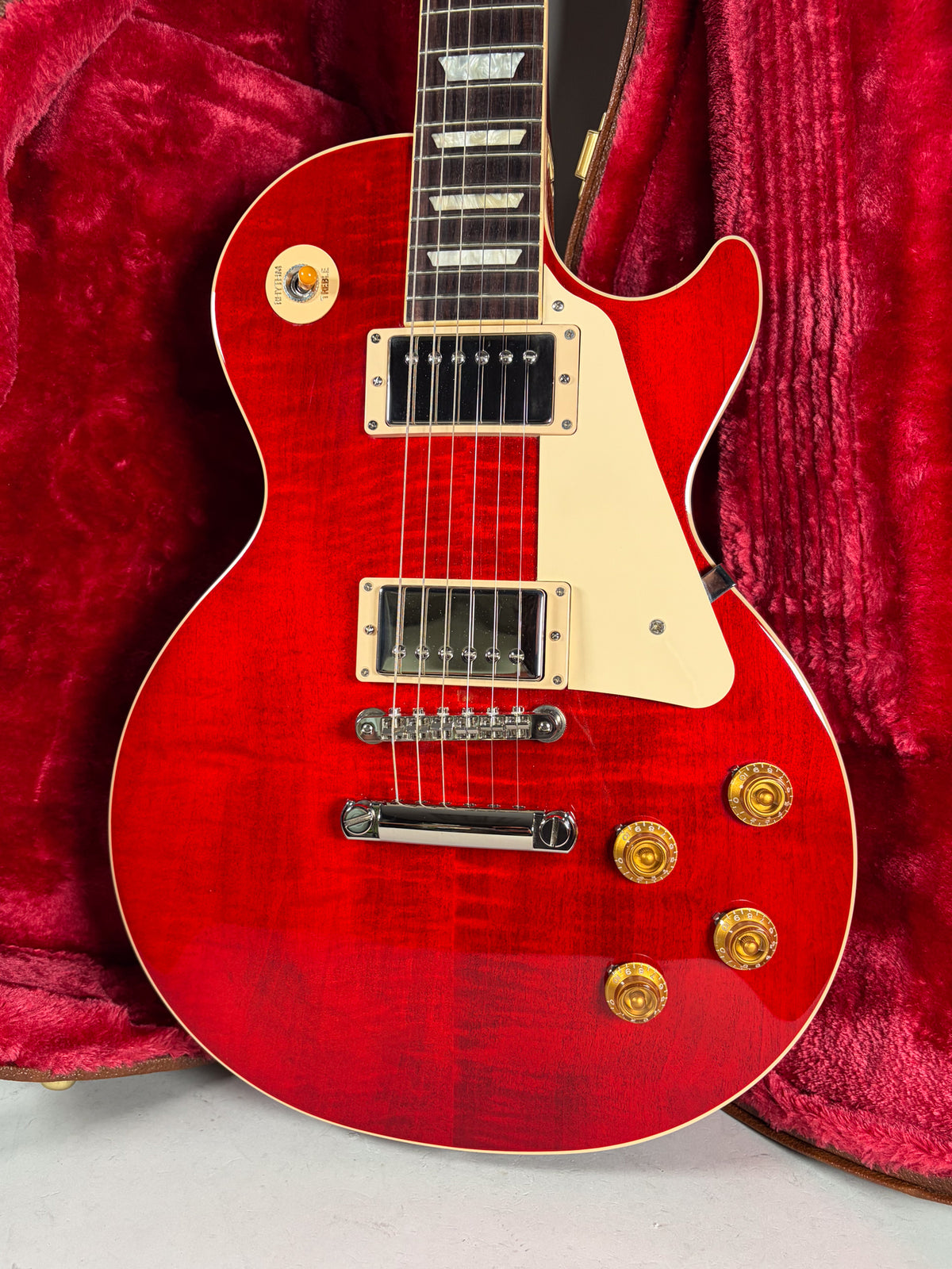 A red flame top Gibson Les Paul Standard 50's electric guitar with cream binding, pickguard, and gold knobs, resting in a red velvet case.