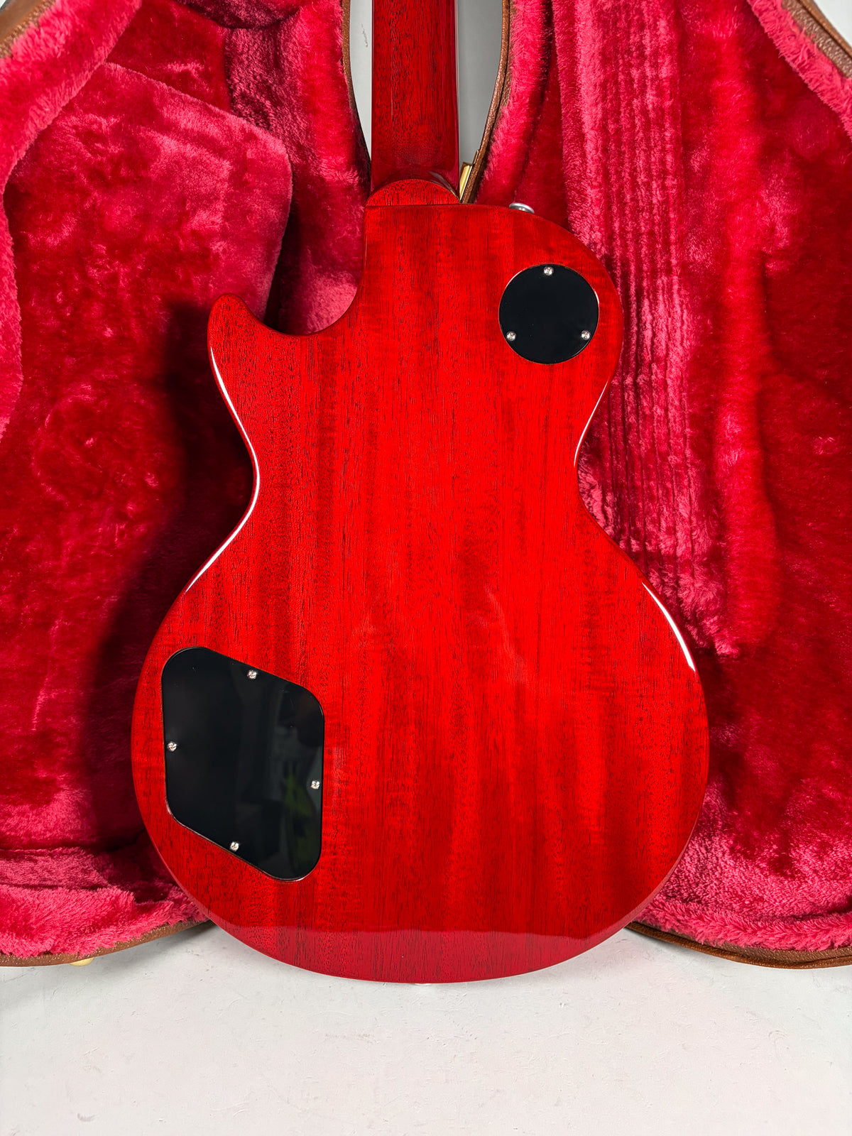 The back of a cherry red Les Paul, showing the mahogany grain, two black control cavity covers, and a red plush case.