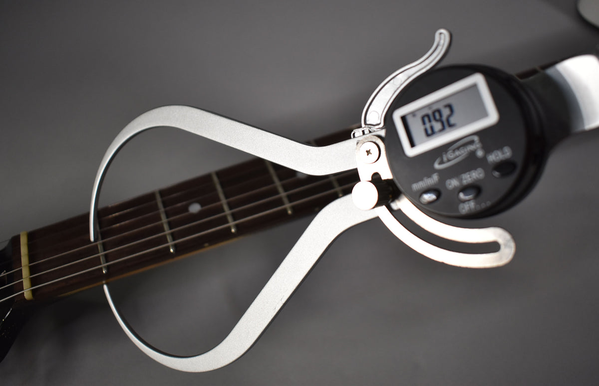 A digital caliper measures the guitar's fretboard, showing "0.92" on its screen, with strings and frets visible.