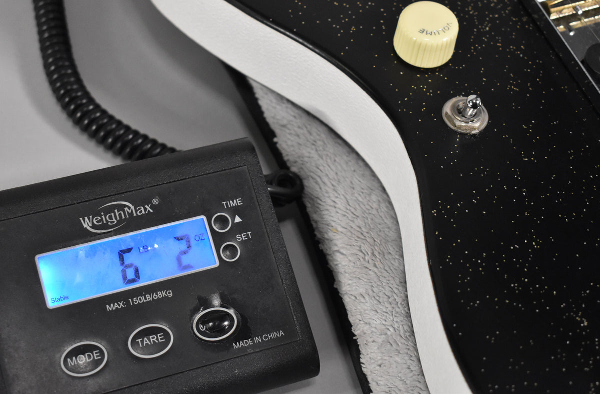 Close-up of the guitar's black sparkle body and cream binding, with a cream volume knob and toggle switch, next to a scale displaying "6 LBS 2 OZ".