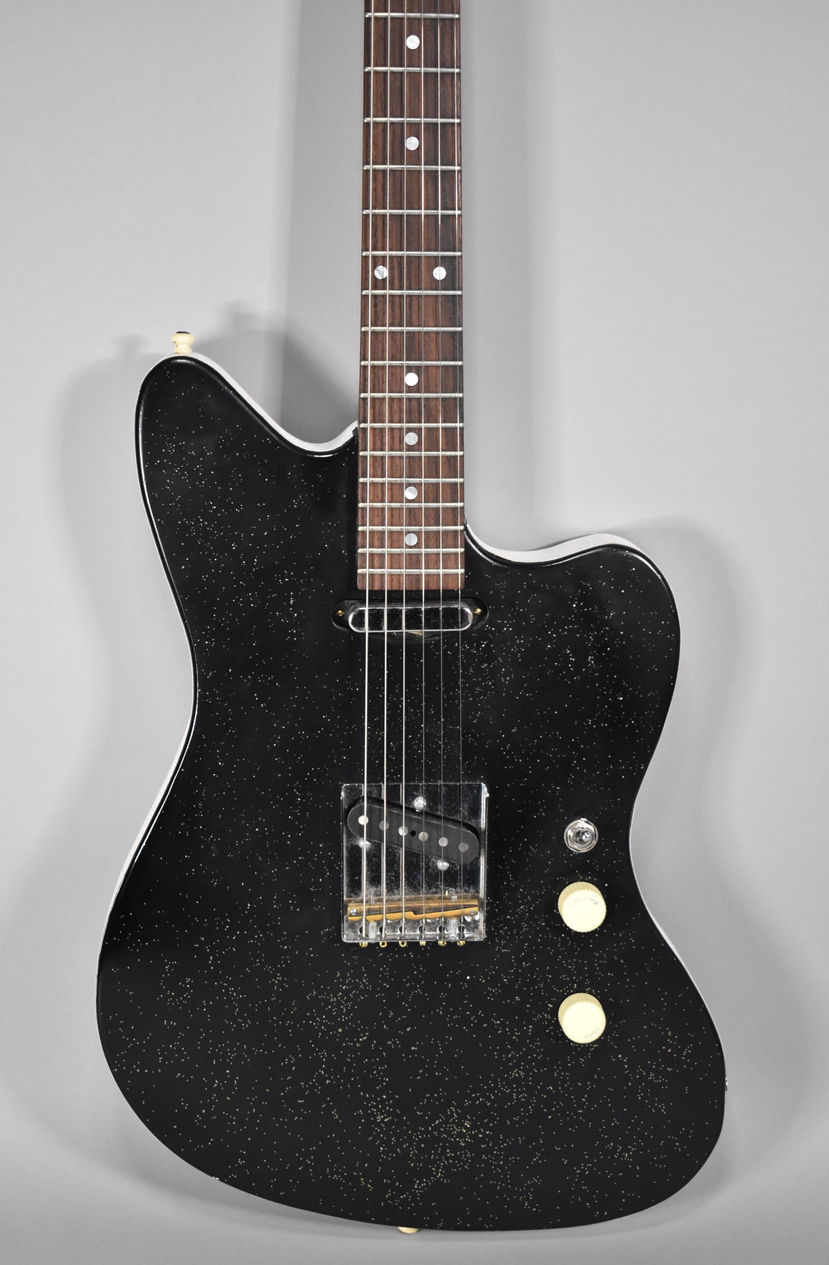 Black sparkle electric guitar, angled from above, showing the body, bridge pickup, two cream control knobs, and part of the neck.