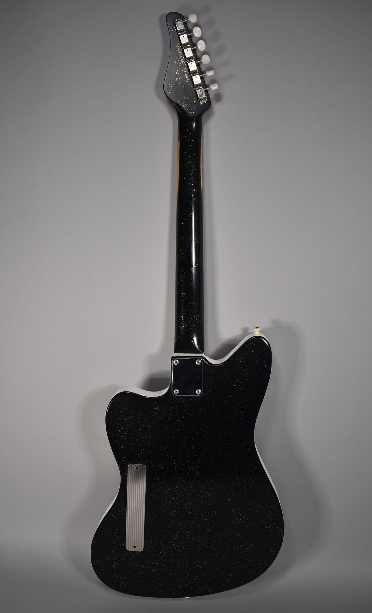 Rear view of a black sparkle guitar with a white binding, a ribbed metal plate, and six white tuning pegs.