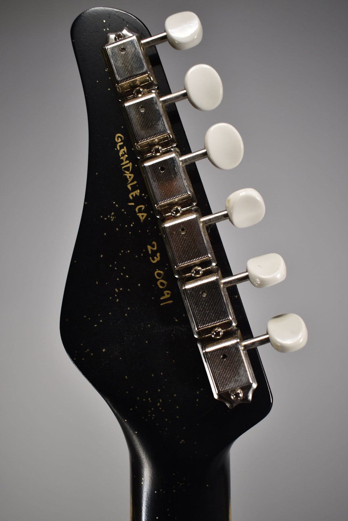 Close-up of a black sparkle guitar headstock with six white tuning pegs and "GLENDALE, CA 23 0091" in gold script.