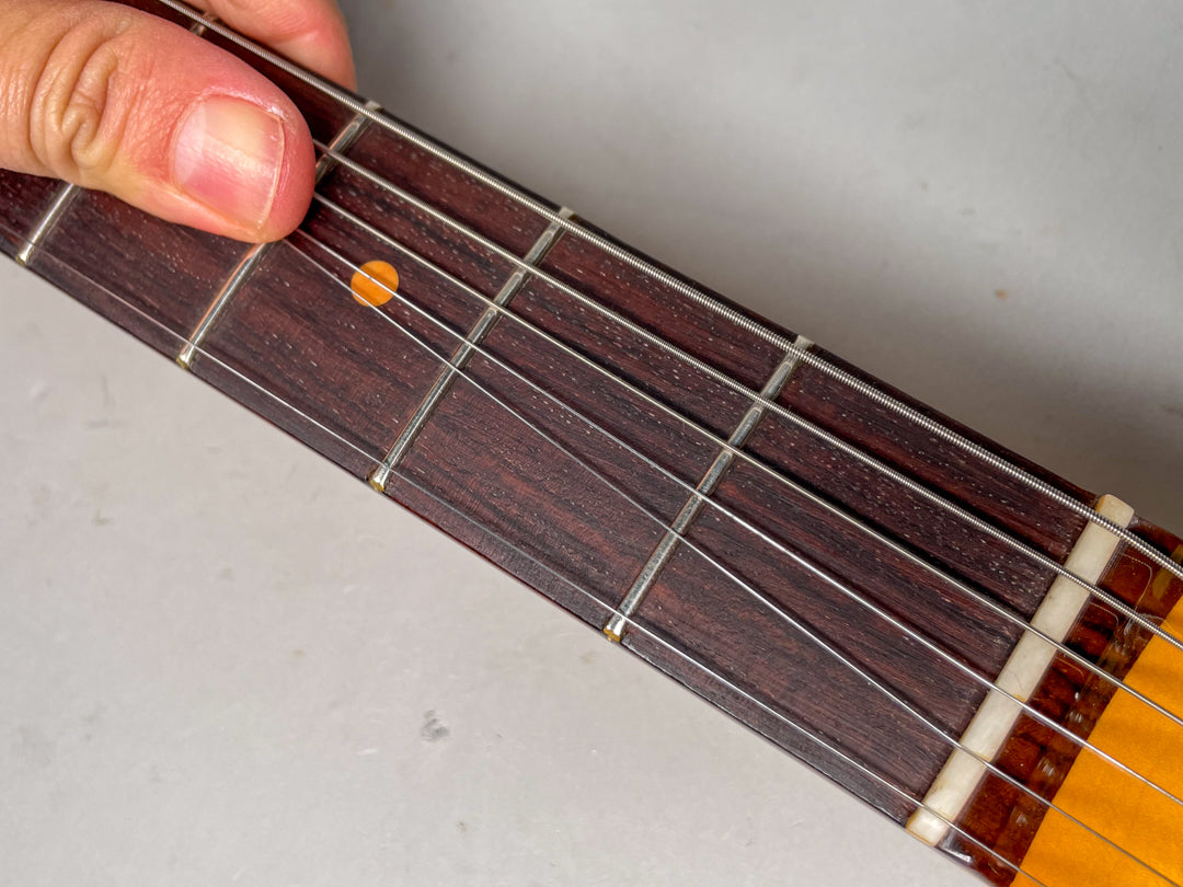 Close-up of the dark rosewood fretboard with a hand pressing the third fret, showing the nut and strings.