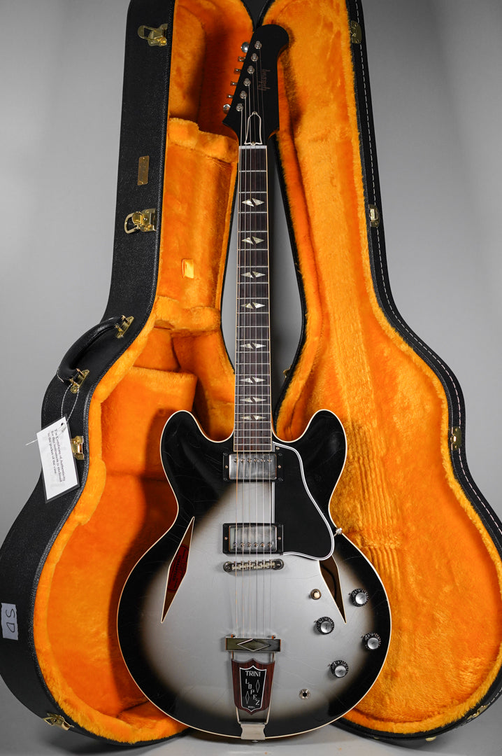 Full view of a silverburst semi-hollow electric guitar with trapezoid inlays, chrome hardware, and four black control knobs, resting in an open orange-lined hard case.